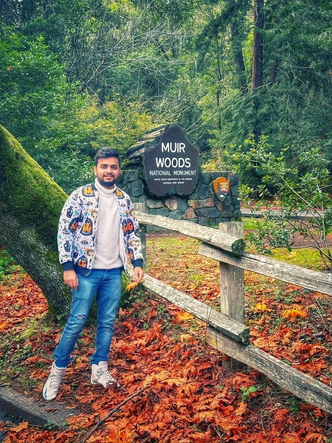 Akshay at the Muir Woods National Monument sign, among redwoods and autumn leaves